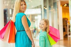 nos encanta ir de compras juntas alegre madre e hija de cabello rubio sosteniendo bolsas de compras y mirando por encima del hombro mientras estamos de pie en el centro comercial foto