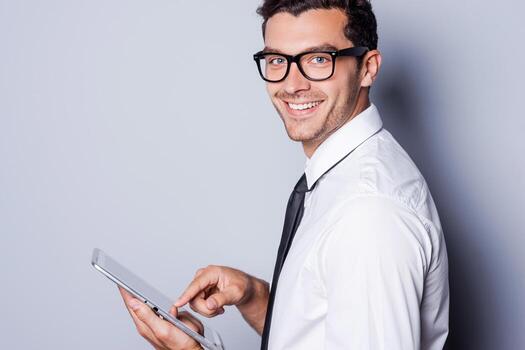 Making business easier. Side view of confident young man in shirt and tie working on digital tablet and smiling while standing against grey background photo