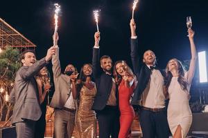 grupo de personas hermosas en ropa formal sosteniendo bengalas y sonriendo mientras pasan tiempo en una fiesta al aire libre foto