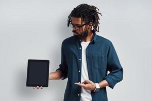Charming young African man in eyewear pointing copy space using digital tablet while standing against grey background photo