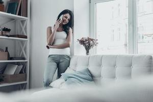 So good to hear from you Attractive young woman talking on the mobile phone and smiling while leaning on the wall at home photo