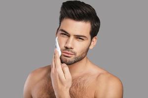 Enjoying morning routine. Handsome young man applying shaving cream and looking at camera while standing against grey background photo