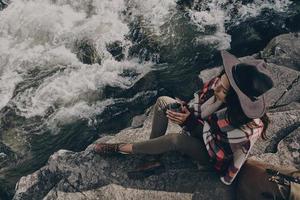 Quite contemplation. Top view of beautiful young woman covered with blanket holding a mug while sitting near the river in mountains photo