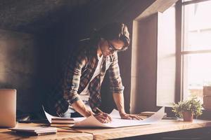 Architect at work. Confident young man in casual wear sketching on blueprint while standing near wooden desk in creative office photo