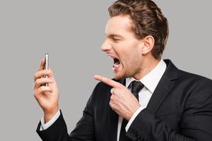 Angry businessman. Furious young man in formalwear holding mobile phone and shouting at it while standing against grey background photo