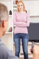 Stop watching TV Rear view of man with remote control watching TV while angry woman standing in front of him and keeping arms crossed photo