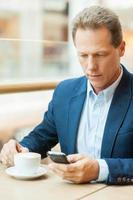 Taking time for coffee break. Confident mature man in formalwear drinking coffee and typing a message on mobile phone while sitting in restaurant photo
