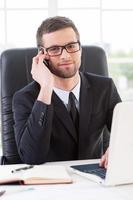 Businessman at work. Confident young man in formalwear talking on the mobile phone and looking at camera while sitting at his working place photo