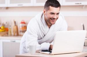 domingo por la mañana sin preocupaciones. un joven apuesto con albornoz trabajando en una laptop mientras está de pie en la cocina foto