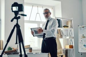 hombre maduro seguro de sí mismo con camisa elegante y corbata que muestra el gráfico y comparte la experiencia empresarial mientras hace videos en las redes sociales foto