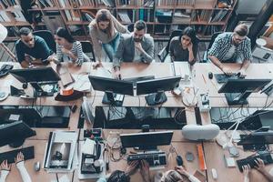 Busy working in office. Top view of group of young business people in smart casual wear working together while sitting at the large office desk photo