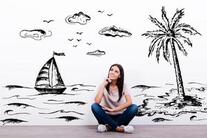 Dreaming about vacation. Cheerful young woman smiling while sitting on the floor with illustration in the background photo
