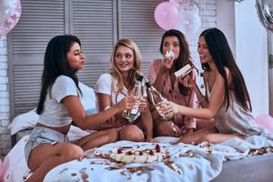 buena fiesta. cuatro hermosas mujeres jóvenes en pijama comiendo pastel y bebiendo champán mientras tienen una fiesta de pijamas en el dormitorio foto