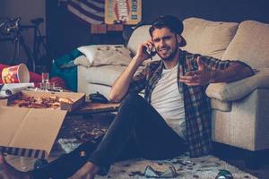 Handsome young man talking on mobile phone and gesturing while sitting on the floor in messy room after party photo