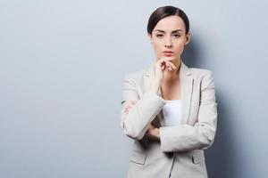 Thoughtful businesswoman. Confident young businesswoman keeping arms crossed and looking at camera while standing against grey background photo