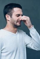 Awful smell Frustrated young man expressing negativity and covering nose with fingers while standing against grey background photo