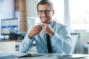 Coffee break. Cheerful young businessman in formalwear holding cup of coffeeand looking at camera while sitting at his working place photo