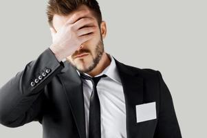 Failed management. Frustrated young man in formalwear covering face with hand while standing against grey background photo