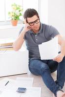 Home finances. Concentrated young man looking at the paper and holding hand on chin while sitting on the floor at his apartment photo