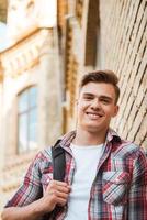 Happy student. Low angle view of handsome young man carrying backpack on one shoulder and smiling while leaning at the brick wall photo