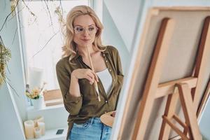 Attractive young blond hair woman painting in art studio and looking thoughtful photo