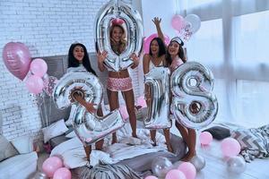 se acerca el año nuevo cuatro atractivas jóvenes sonrientes en pijama sosteniendo globos mientras están de pie en el dormitorio en casa foto