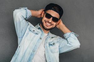 alegre y moderno. un joven alegre ajustando su gorra de béisbol y mirando a la cámara con una sonrisa mientras se enfrenta a un fondo gris foto