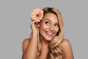 Cute young woman adjusting flower in her hair and smiling while standing against grey background photo