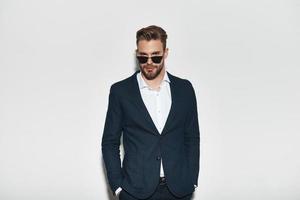 True elegance. Handsome young man in full suit keeping hands in pockets and looking at camera while standing against grey background photo