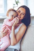 Best hug ever. Cheerful beautiful young woman holding baby girl in her hands and looking at camera with smile while sitting on the couch at home photo