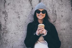 Her smile can melt your heart. Beautiful young woman in sunglasses holding coffee cup and looking at camera with smile while standing against concrete wall outdoors photo