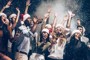 They love Christmas. Group of beautiful young people in Santa hats throwing colorful confetti and looking happy photo