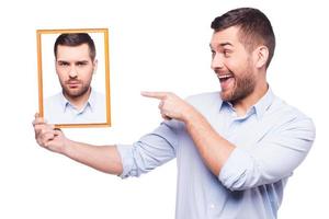 dame una sonrisa apuesto joven con camisa sosteniendo una foto de sí mismo con la cara molesta y señalándola con una sonrisa mientras está de pie contra un fondo blanco