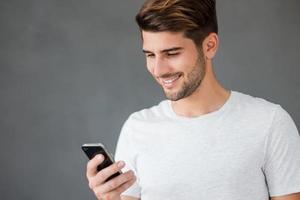 Texting to friend. Happy young man holding mobile phone and smiling while standing against grey background photo