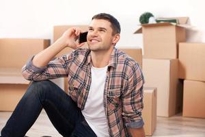 Calling from a new house. Cheerful young man sitting on the floor and talking on the mobile phone while cardboard boxes laying on the background photo