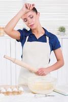 cansado de cocinar. mujer joven cansada en delantal sosteniendo un rodillo y tocándose la frente con la mano mientras estaba de pie en una cocina foto