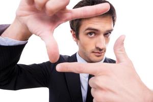 Focusing at you. Wide-angle image of young man in formalwear gesturing finger frame and looking through it while standing isolated on white background photo