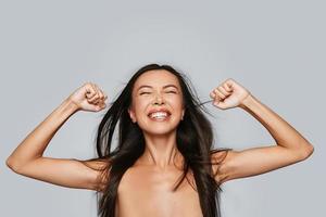 Winning. Beautiful young Asian woman keeping eyes closed and gesturing while standing against grey background photo