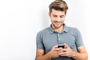 Always in touch with friends. Handsome young man holding mobile phone and looking at it with smile while leaning at the wall photo