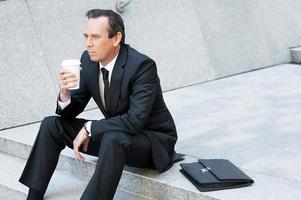 Relaxing after hard working day. Thoughtful mature man in formalwear holding coffee cup and looking away while sitting at the staircase photo