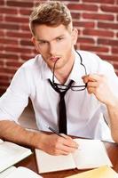 I need some fresh ideas. Top view of thoughtful young man in shirt and tie writing something in note pad and looking away while sitting at his working place photo