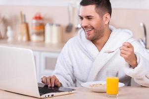 trabajando durante el desayuno. un joven apuesto que toma un desayuno saludable y trabaja en una laptop mientras está sentado en la cocina foto