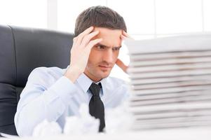 That is too much for me Depressed young man in shirt and tie holding head in hands while sitting at the table with stack of paperwork on it photo