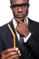 I need a new one. Thoughtful young African man in formalwear holding pencil and looking at it while holding hand on chin and standing isolated on white background photo