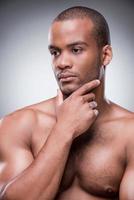 Lost in thoughts. Portrait of young African man holding hand on chin while standing against black background photo