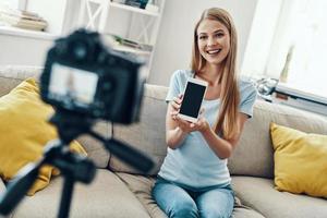 hermosa mujer joven sonriendo y señalando espacio de copia en su teléfono inteligente mientras hace videos de medios sociales en casa foto