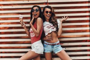 Enjoying carefree time. Two attractive young women smiling and holding ice cream while standing against the wooden wall outdoors photo