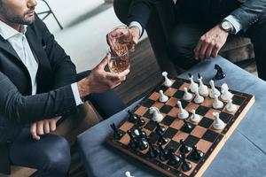 Great way to relax. Close up top view of young men in full suits playing chess and toasting each other while sitting indoors photo