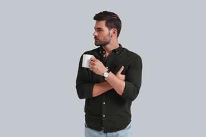 Taking time to think. Serious young man holding a cup and looking away while standing against grey background photo