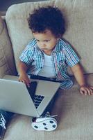 Multitasking baby. High angle view of little African baby boy using his laptop and smartphone while sitting on the couch at home photo